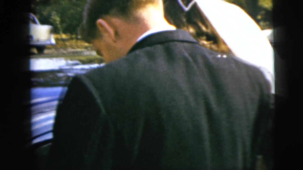 Groom in a dark suit stands beside a blue car as a woman in white assists nearby.
