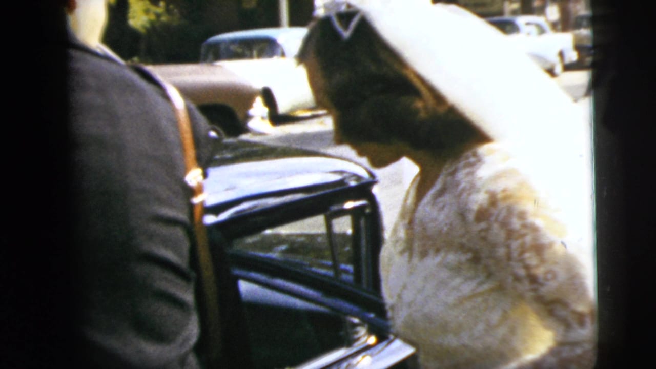 In Moline, Illinois, a bride in a veil and lace gown stands beside a dark car during a 1962 wedding departure.