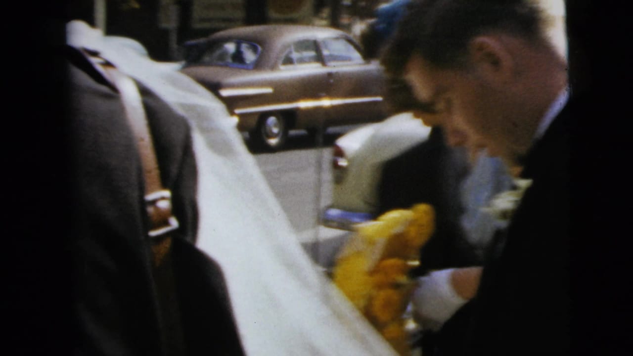 A suited man assists the bride with fabric from her gown or veil beside a car during the Moline wedding departure.