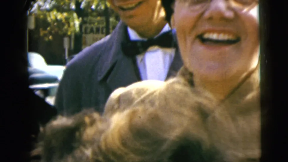 A laughing woman fills the frame beside the car window while a suited man in a bow tie stands behind her on the street.