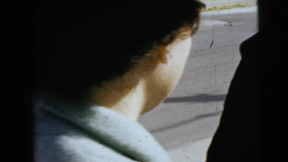 The camera catches the back and side of the woman's head and pale coat near the car window, with street pavement visible beyond.