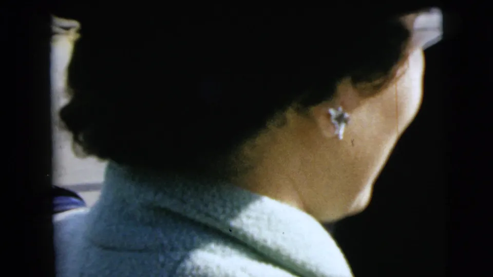 The woman turns into profile near the car window, her earring and pale coat visible in the 1962 Moline home-movie frame.