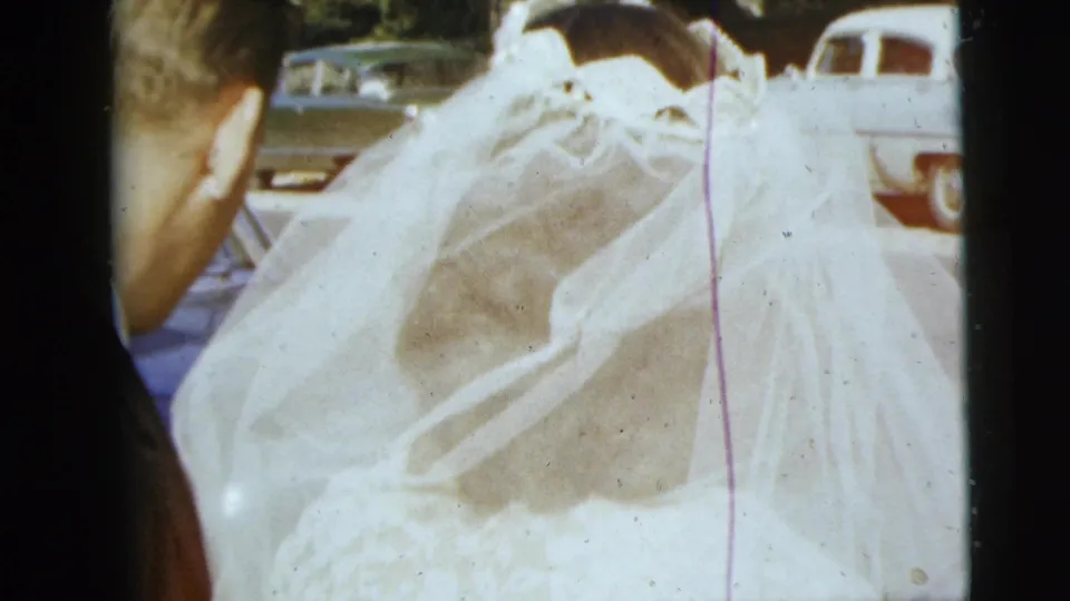 In Moline, Illinois home-movie footage from 1962, the bride's veil fills the frame as the couple stands near parked cars outdoors.