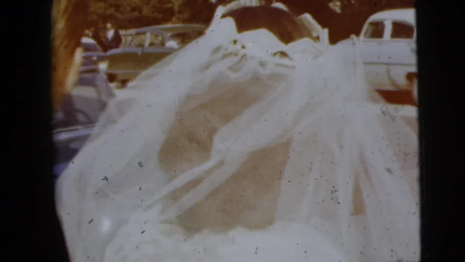 The bride's veil and gown dominate the frame while the couple stands outdoors near mid-century parked cars in 1962 home-movie footage.