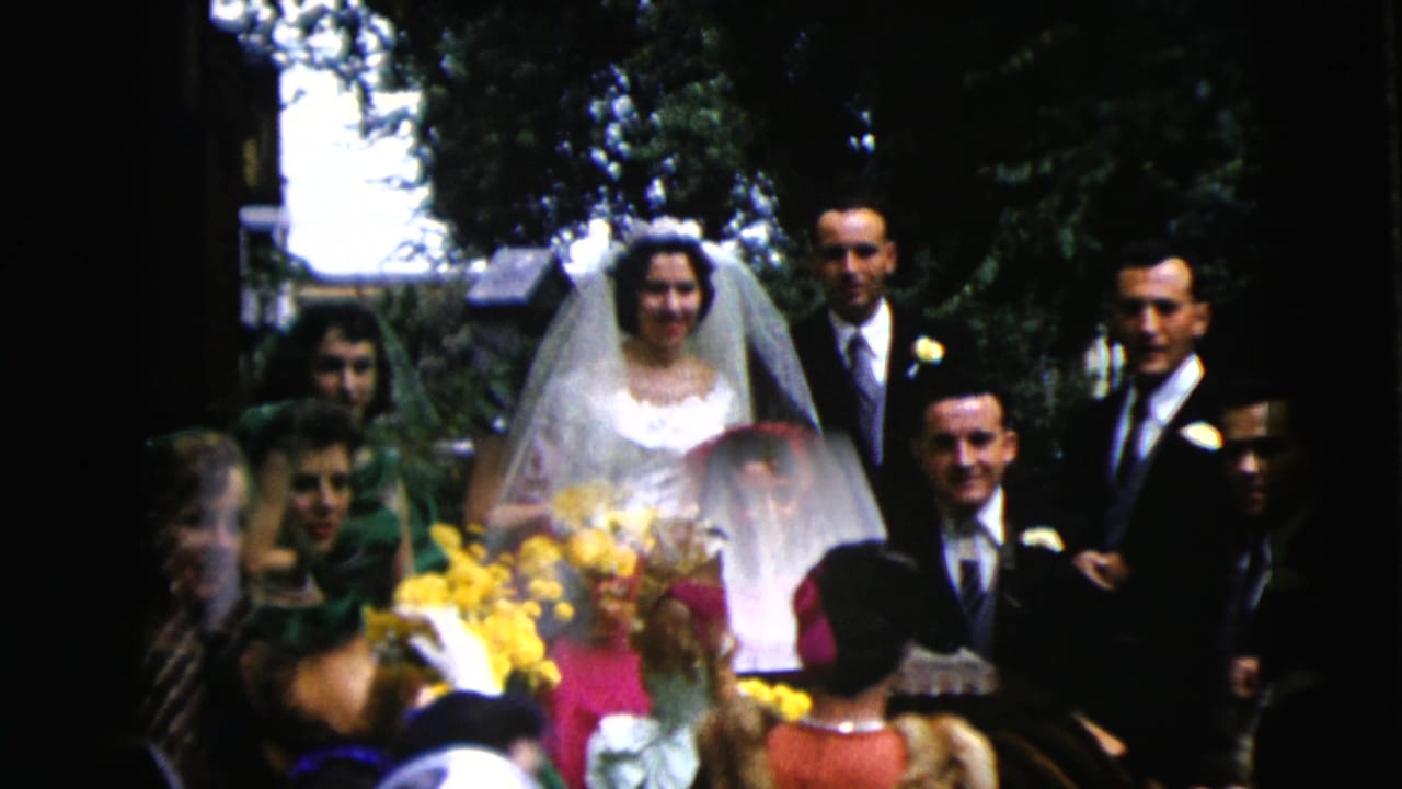 Bride with veil and yellow bouquet stands with groom and wedding party among guests outdoors.
