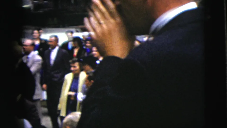 1962 Moline wedding home movie frame with formal guests gathered near the ceremony, partly obscured by a close foreground figure.