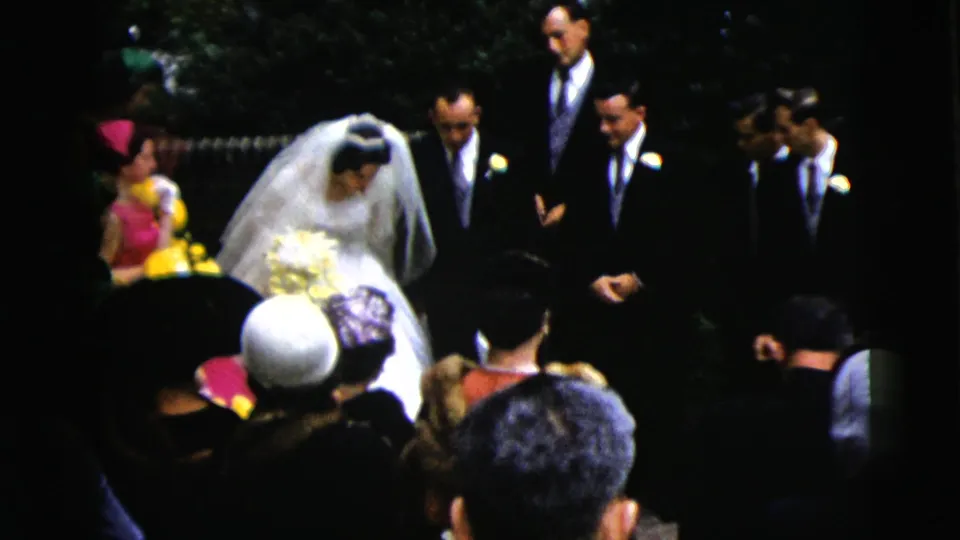 In this 1962 Moline wedding home movie frame, the bride in a white veil approaches groomsmen as guests watch from the aisle.