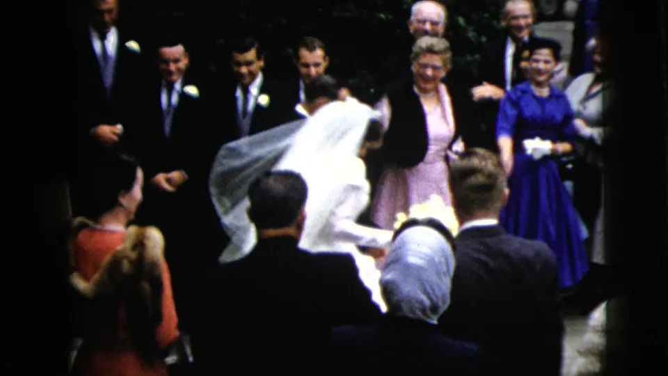 The bride moves forward beside the wedding party and nearby guests, with older attendees and colorful dresses visible in the ceremony space.