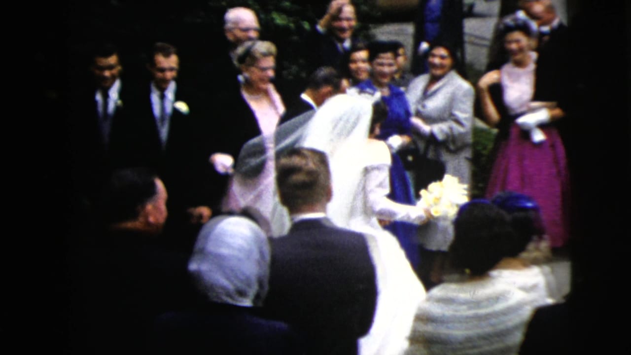 Bride in a white gown approaches a gathered wedding crowd outside.