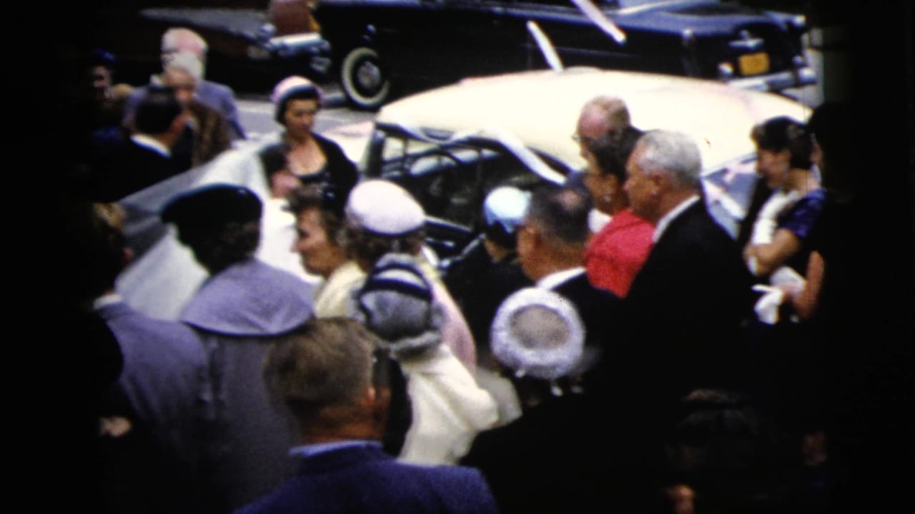 Wedding guests surround a white car as the couple approaches.