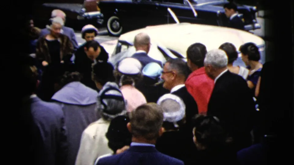 A dense group of wedding guests stands around a white sedan while the couple prepares to enter or approach the car.
