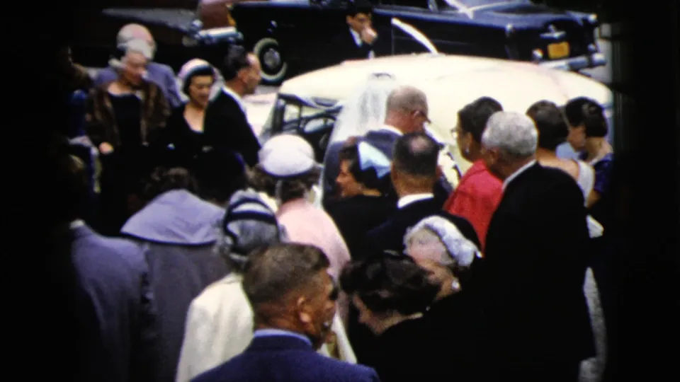 The bride's veil appears beside the white sedan as guests in formal clothing continue to watch the wedding send-off.