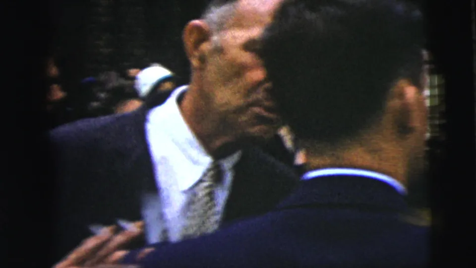 Close handheld 1962 Moline church-event view with suited men and blurred guests standing in a dense outdoor crowd.