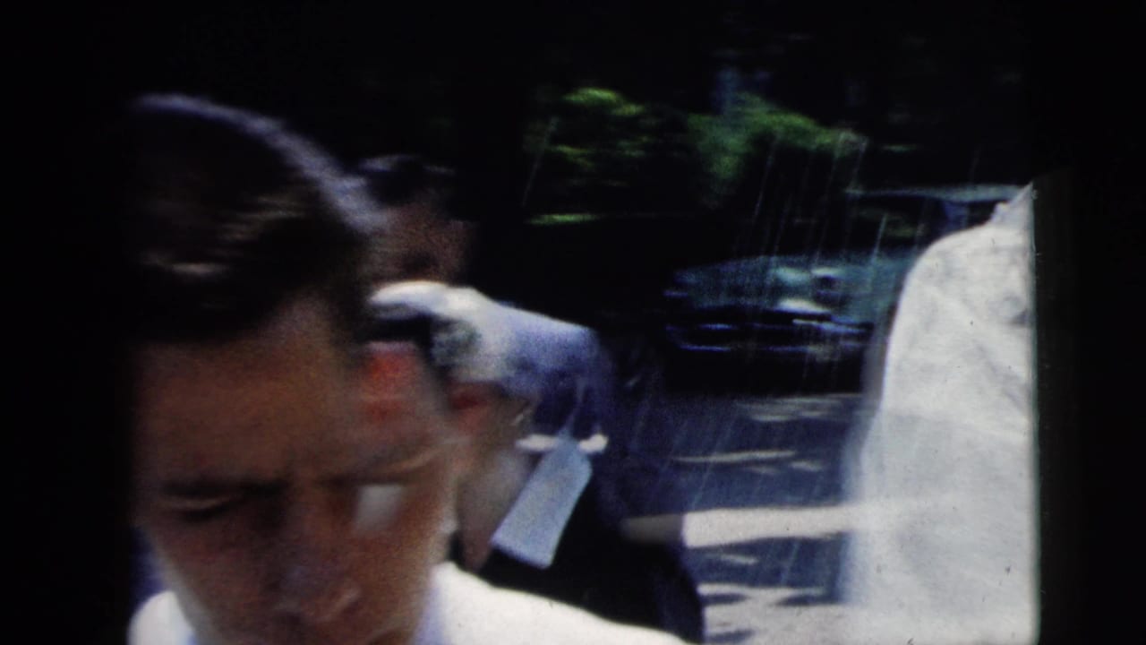 Close handheld 1962 Moline wedding view of a man among guests, with bright sun and a parked car in the background.