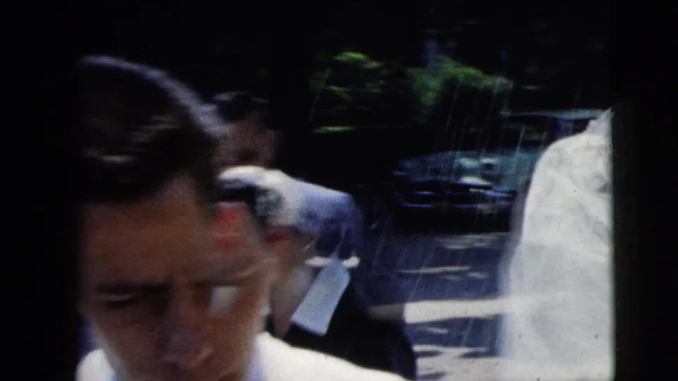 Close handheld 1962 Moline wedding view of a man among guests, with bright sun and a parked car in the background.