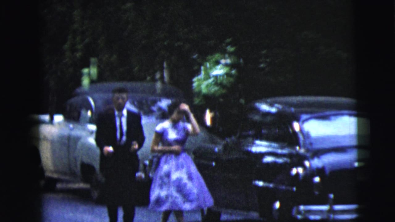 Man in dark suit and woman in purple dress walk beside parked vintage cars.