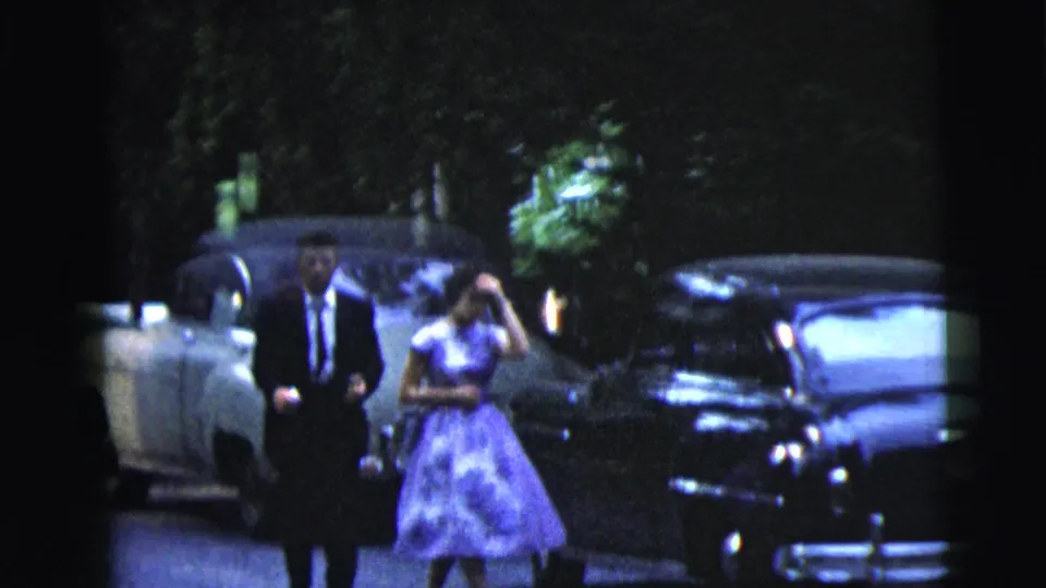 In Moline, Illinois, 1962 home-movie footage, a formally dressed couple walks near parked mid-century cars at night.