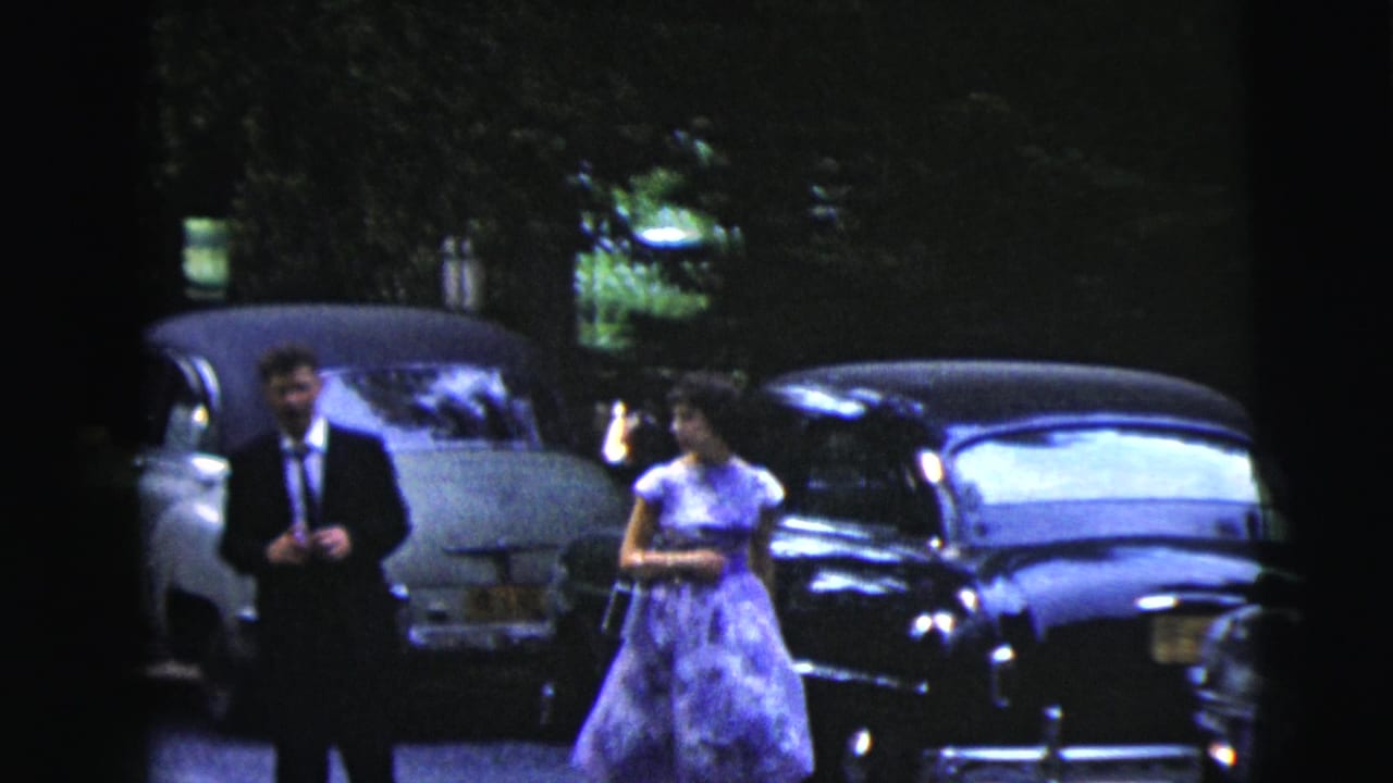 Formal couple stands or walks near a light sedan and dark parked car.