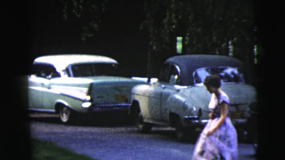 A woman in a purple dress walks across the frame past parked mid-century cars in Moline, Illinois, 1962 footage.