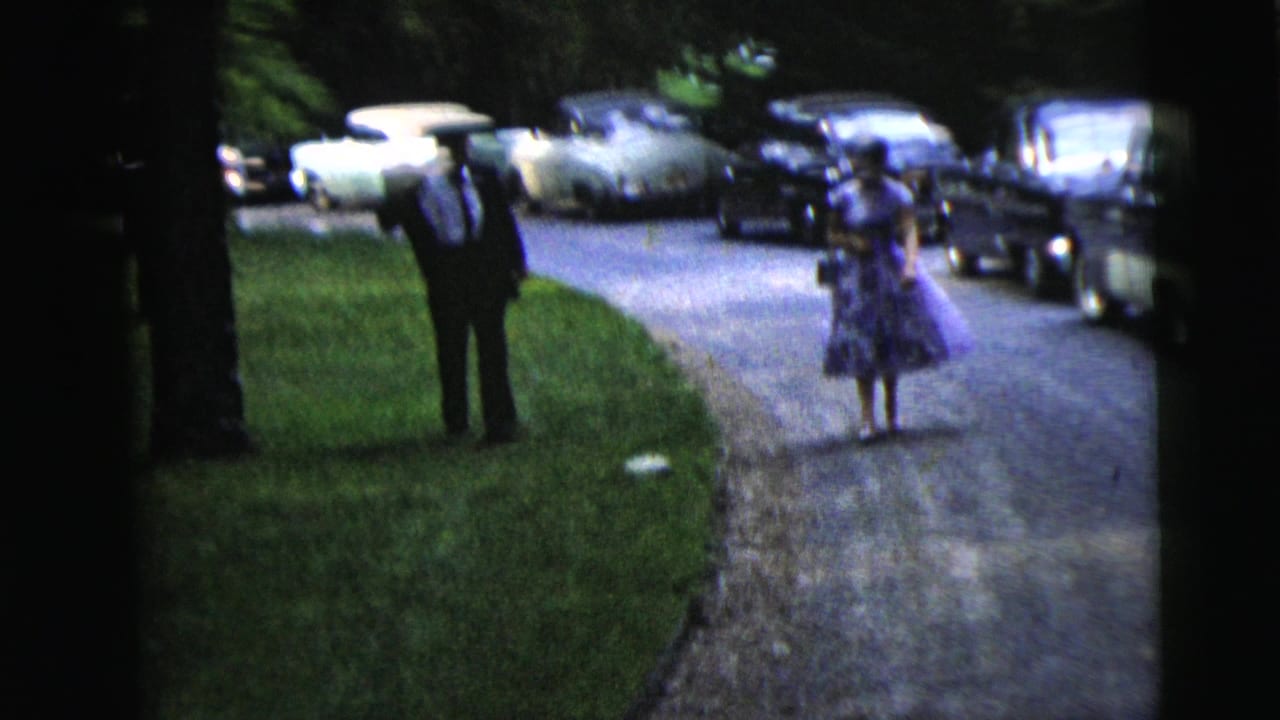 Man and woman walk on a driveway beside parked classic cars and a lawn.
