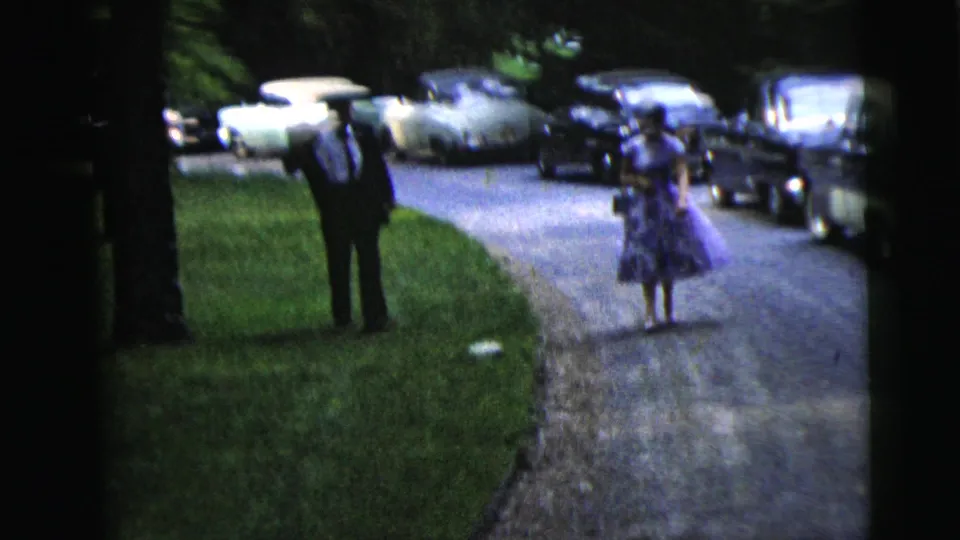 In 1962 Moline, Illinois home-movie footage, a suited man and a woman in a purple floral dress walk along a driveway lined with cars.