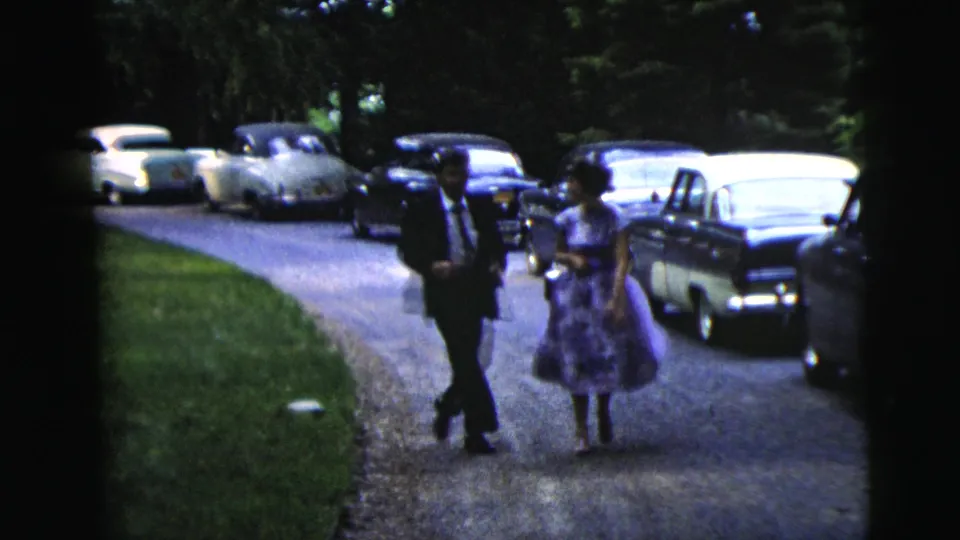 The suited man and woman in a purple floral dress walk side by side toward the camera on a driveway in 1962 Moline, Illinois.