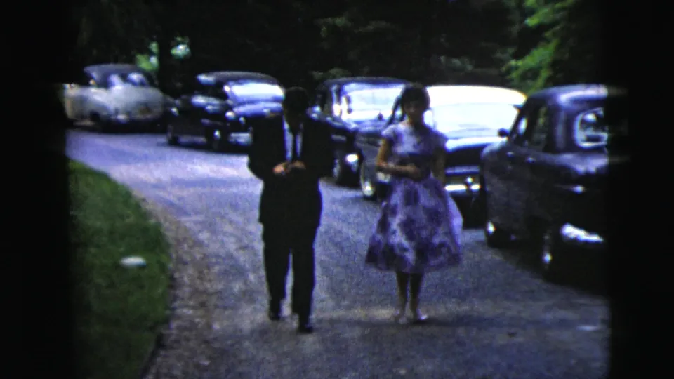 A man in a dark suit and a woman in a purple floral dress move past the camera on a driveway in Moline, Illinois, with classic cars nearby.