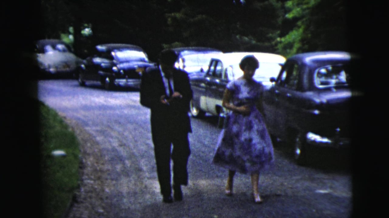 A man in a suit and a woman in a purple dress walk along a gravel road past parked cars.