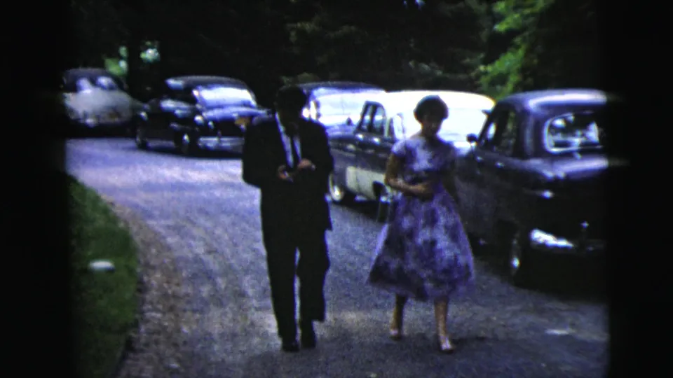In 1962 Moline, Illinois, a suited man and a woman in a purple dress walk toward the camera on a gravel road lined with parked cars.