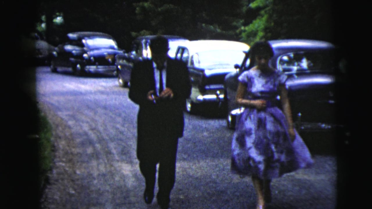 The man in a dark suit and the woman in a purple dress approach the camera along a shaded Moline road in 1962.