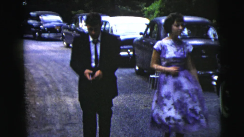 A suited man and a woman carrying a small purse walk side by side near parked cars in 1962 Moline, Illinois.