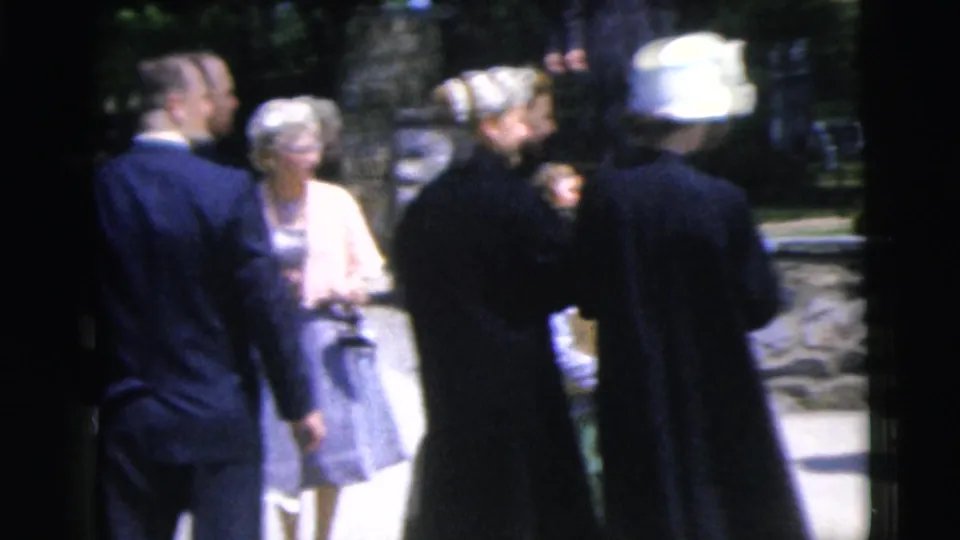 Adults in suits, dresses, and hats gather outdoors in the 1962 Moline home-movie scene.