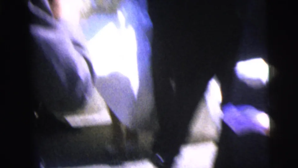 Close, blurred 8mm frame from the wedding scene with figures in formal clothing passing near the camera.