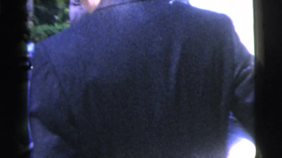 A man in a dark suit fills the foreground while the wedding party remains partly visible in the bright outdoor scene.