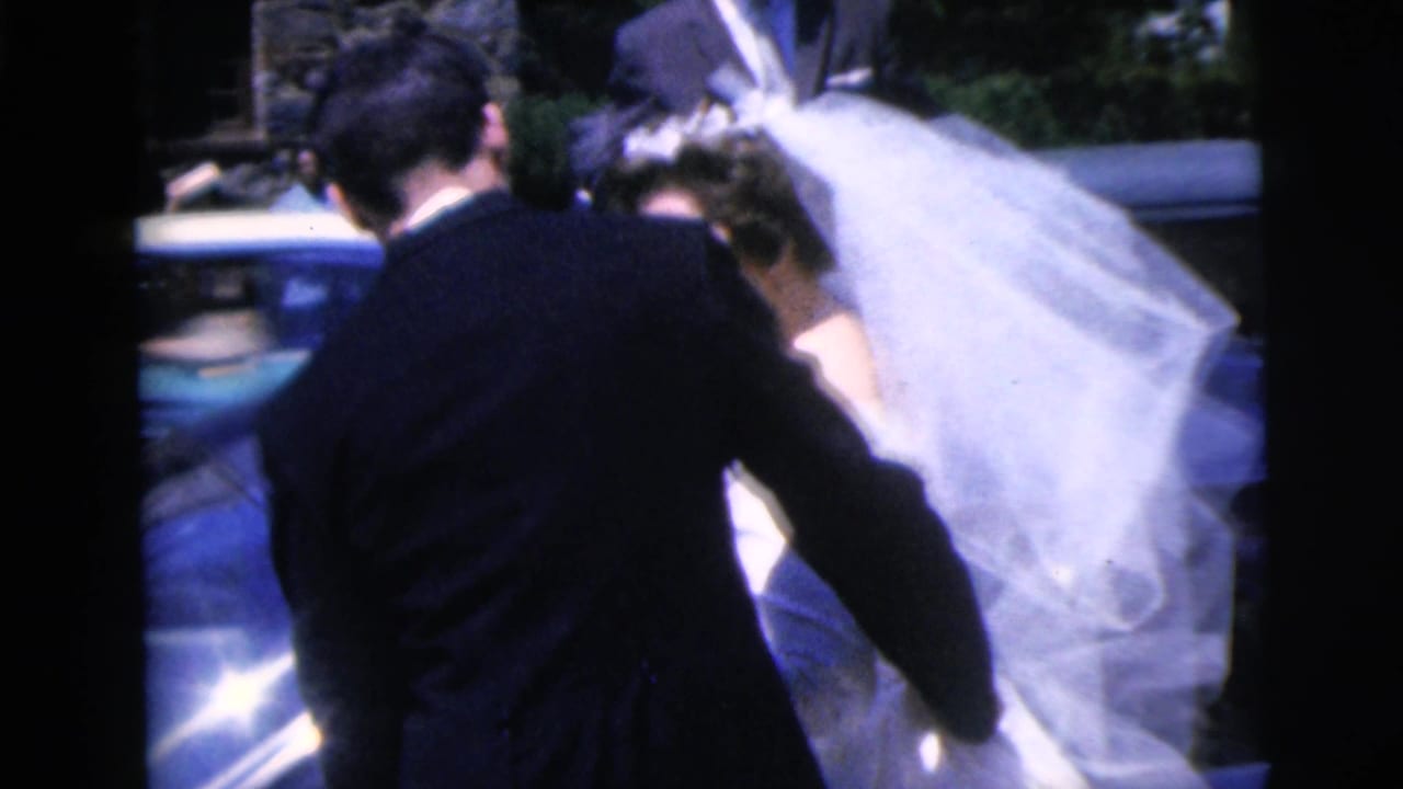 Groom in a dark suit guides a veiled bride beside a blue car.