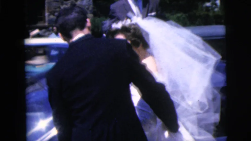 1962 Moline wedding home movie frame showing the groom guiding the veiled bride beside a blue car near a stone building.