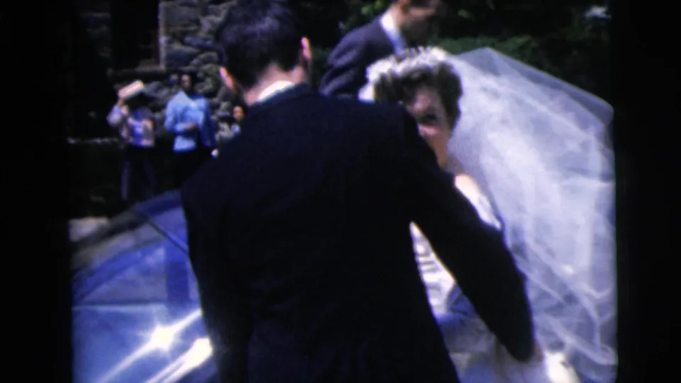 1962 Moline wedding frame with bride and groom beside a blue car while guests watch near the stone building.