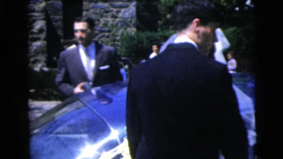 1962 Moline wedding home movie frame showing a suited man near the car as the groom turns toward the bride.