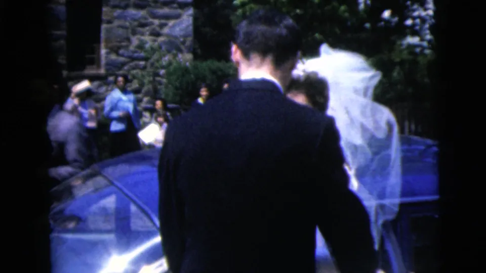 1962 Moline wedding frame with the groom and veiled bride beside a blue car, guests and stonework in the background.