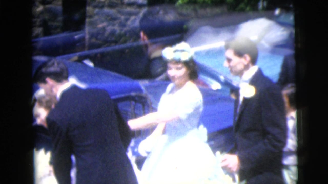 Bride in white gown and floral crown stands near a blue car with men in dark formalwear.