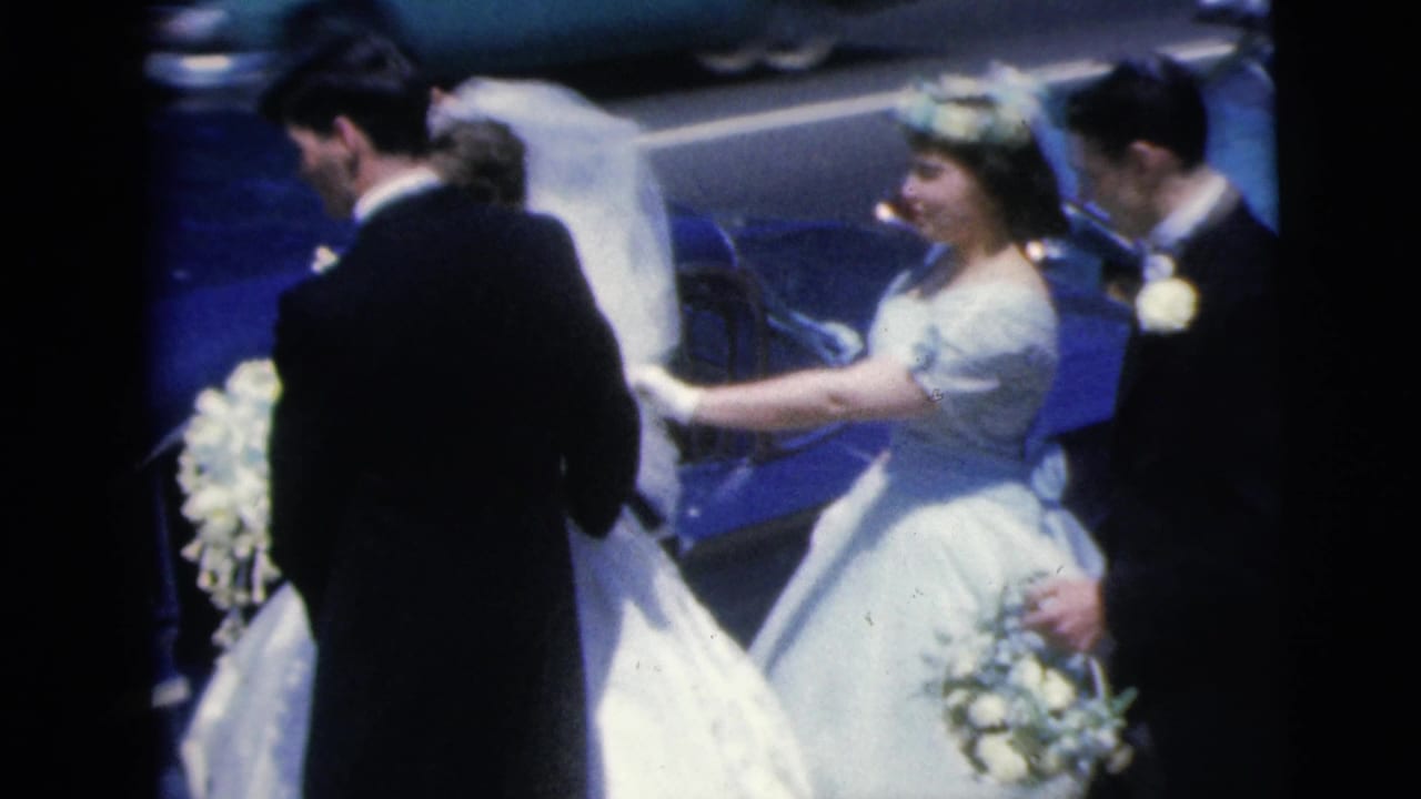 The bride in white stands beside the blue car with her veil visible and attendants in formalwear nearby.