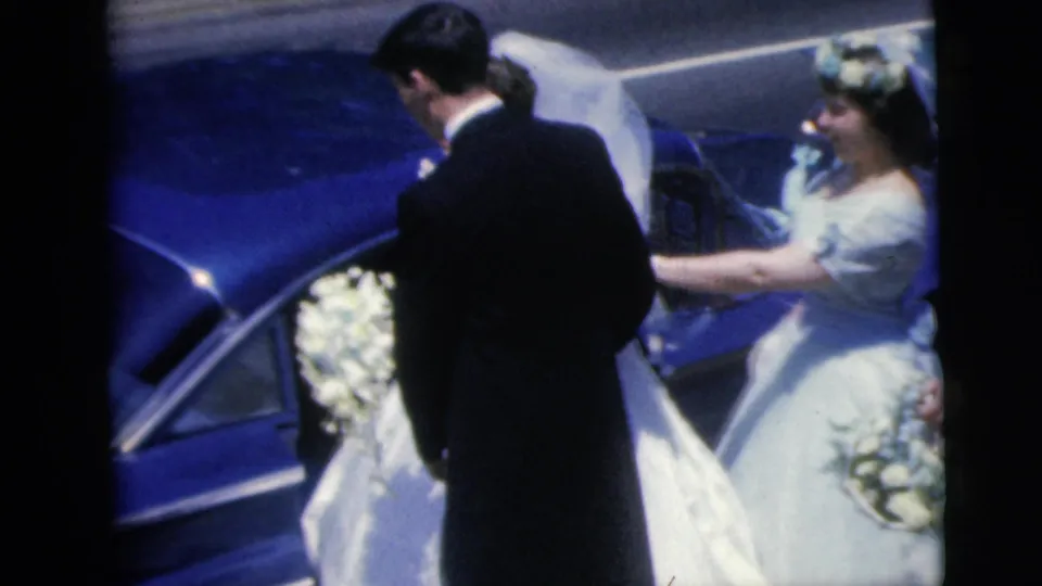 1962 wedding footage shows the bride beside the blue car with a pale-dressed attendant and bouquet just behind her.