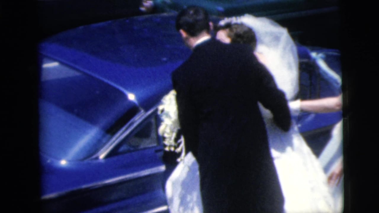 Bride and groom beside a blue car as guests help with the dress and bouquet.