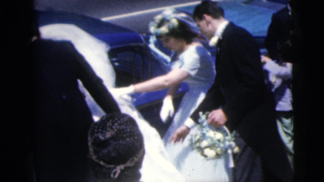 Bride holding flowers is guided near a blue car by the groom and guests.