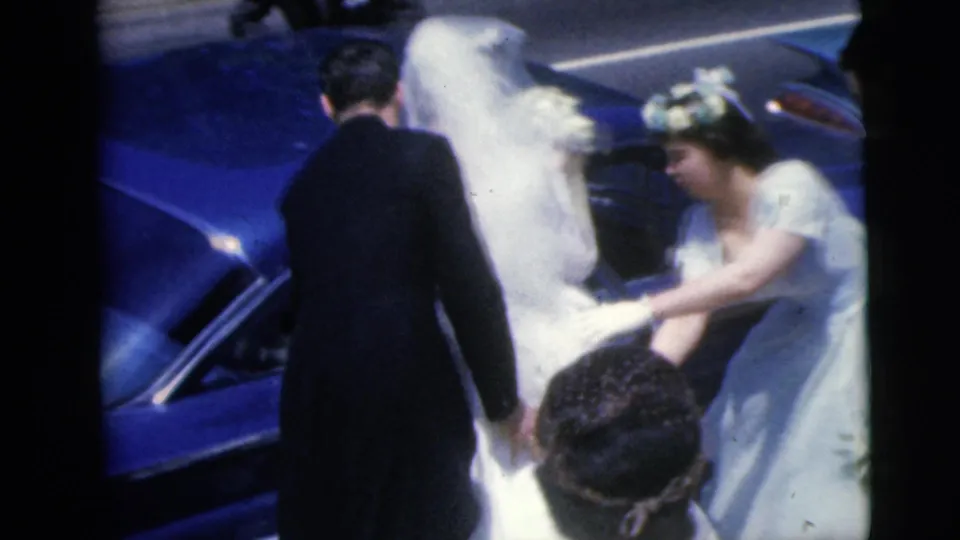 The bride is helped near the side of a blue car during a 1962 Moline wedding, with the groom and guests close around her.