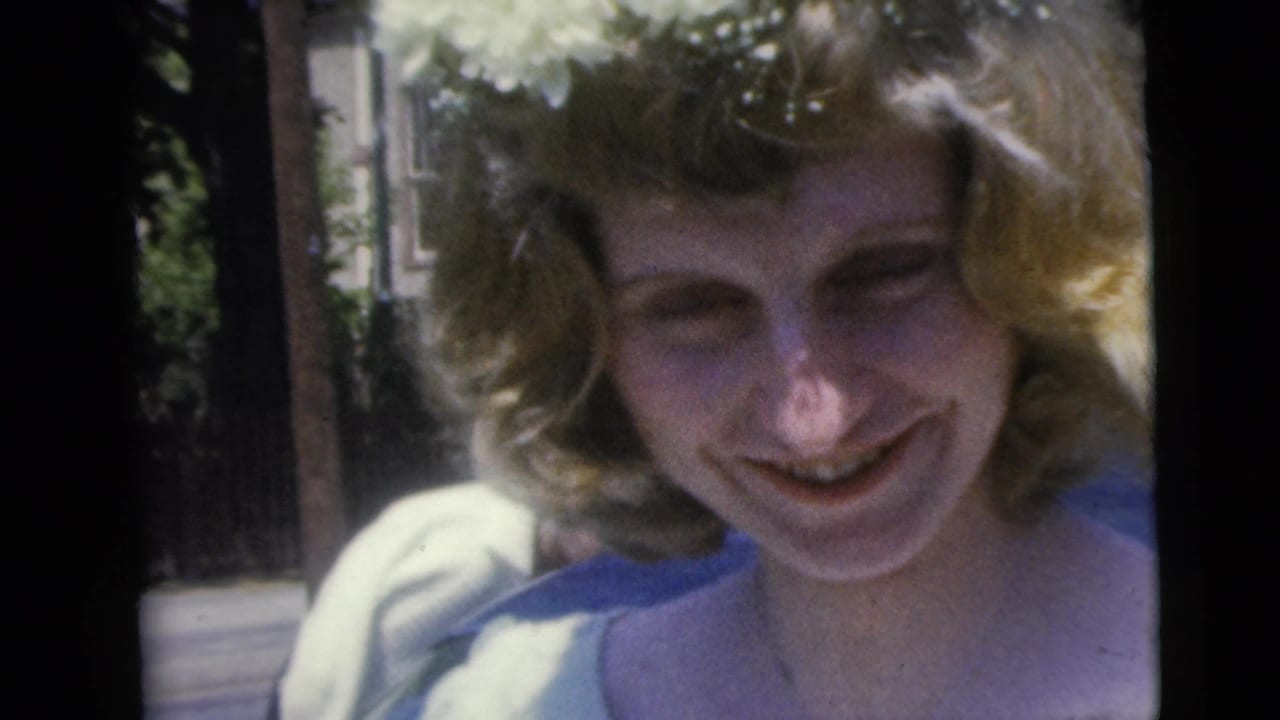 A blonde young woman smiles toward the camera in 1962 Moline, Illinois home-movie footage, with trees and sunlit background behind her.