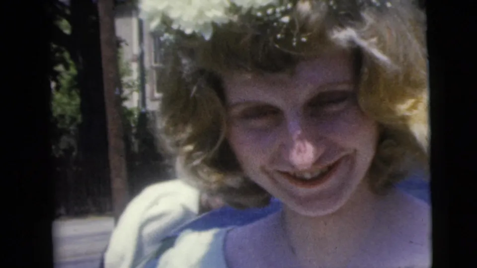 A blonde young woman smiles toward the camera in 1962 Moline, Illinois home-movie footage, with trees and sunlit background behind her.
