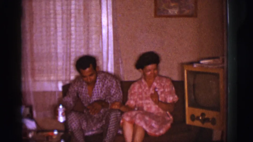 1966 Clarkdale home-movie frame of two adults seated on a small couch in patterned clothing near a wooden television cabinet.