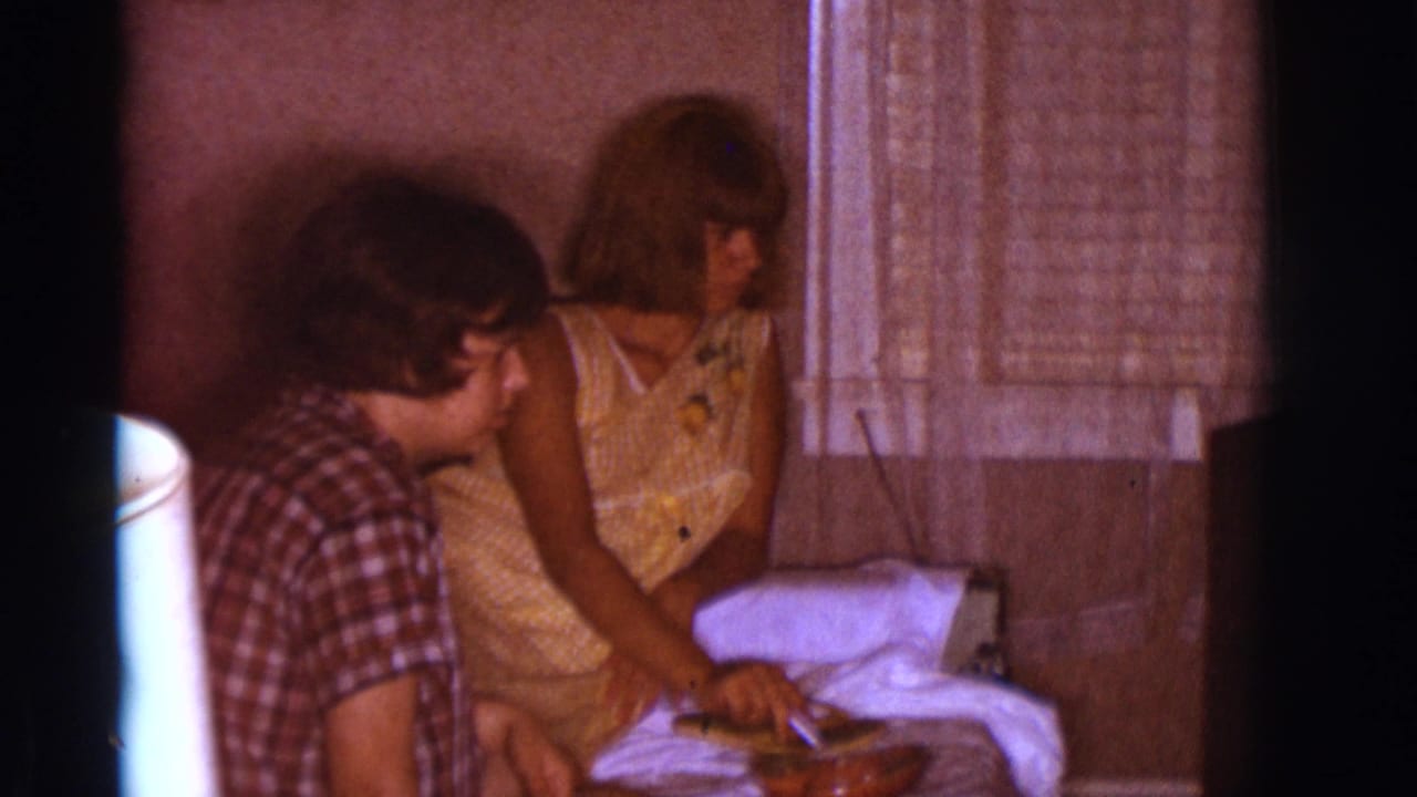 Young man and woman seated indoors beside bedding while petting a small dog.