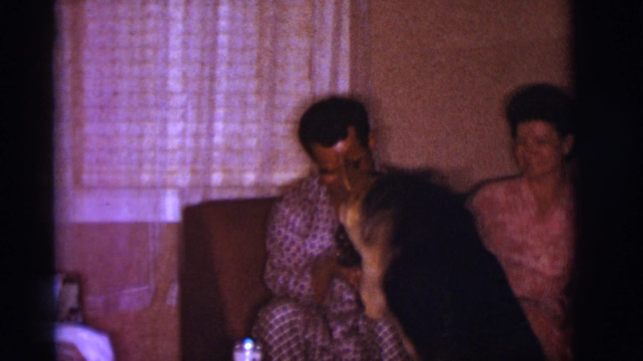 In a Clarkdale, Arizona 1966 home-movie frame, a man and woman sit on a couch while a German Shepherd rises toward the man.